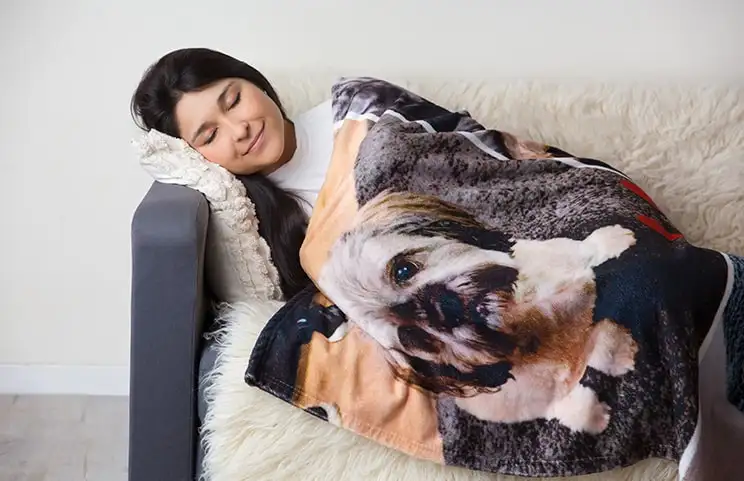 A woman wrapped in a personalised Printerpix mink blanket with a dog design. She is happily asleep on the sofa.